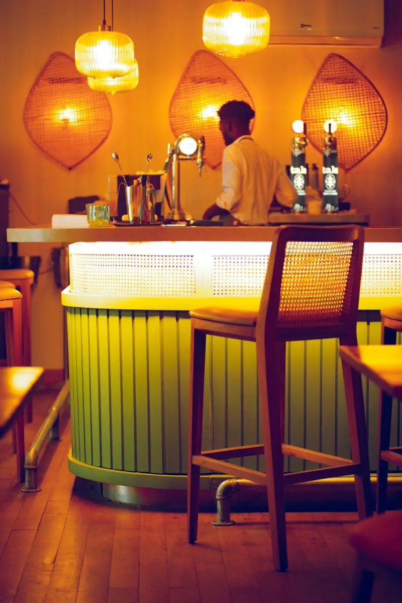 Bar med limegrön front och barstolar med rottingrygg, en bartender under varmt ljus från pendellampor