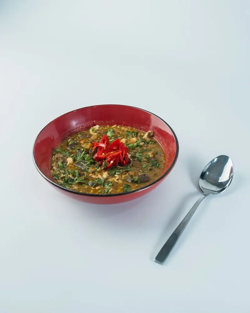 Grön, kryddig soppa med bladgrönsaker och köttbitar, toppad med färsk röd chili i röd skål